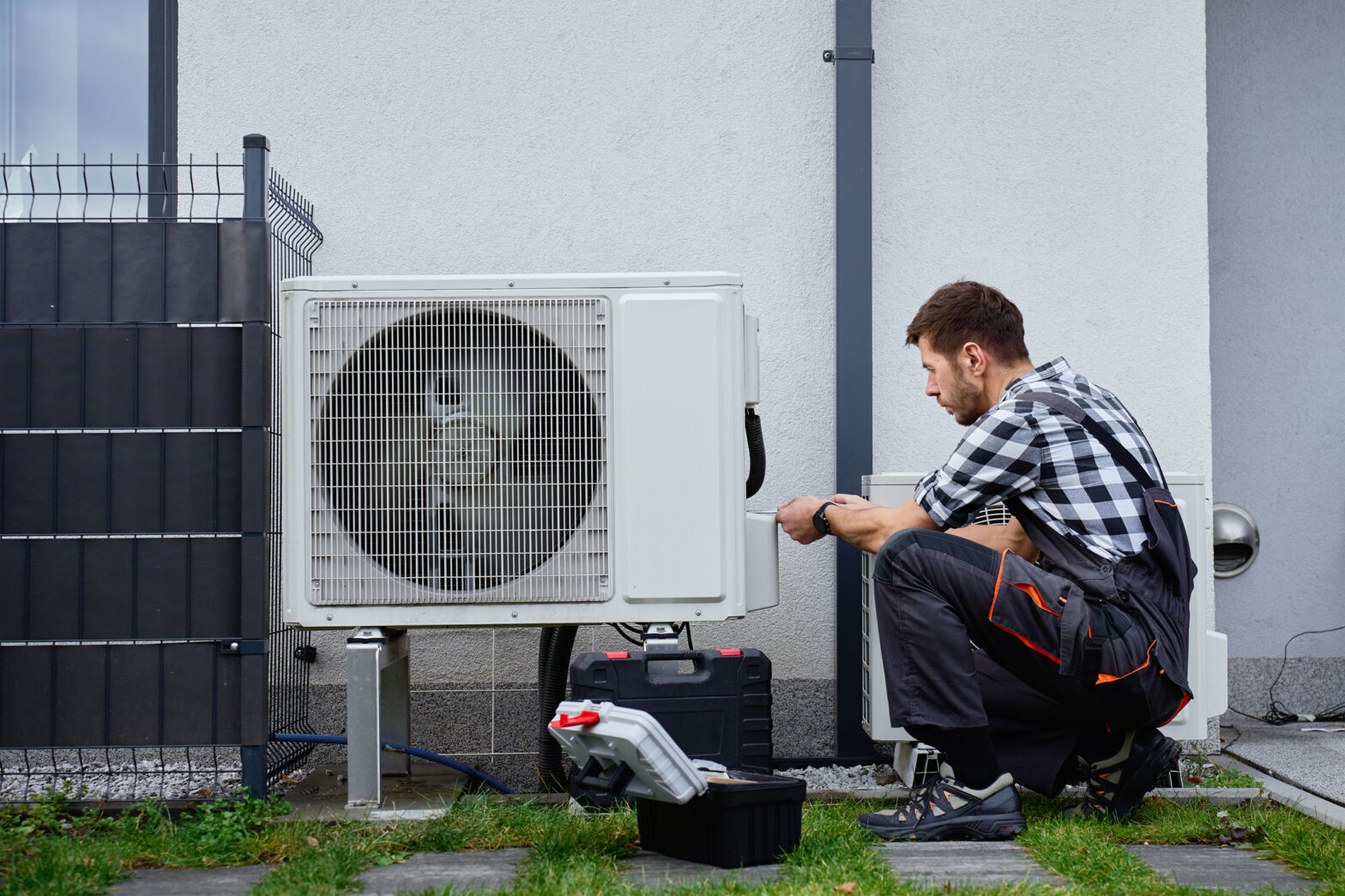 Technician performing maintenance on outdoor heat pump unit near residential building exterior. Man installing air source heat pump, using tools. Concept of HVAC service and heating system repair