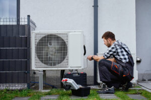 Technician performing maintenance on outdoor heat pump unit near residential building exterior. Man installing air source heat pump, using tools. Concept of HVAC service and heating system repair