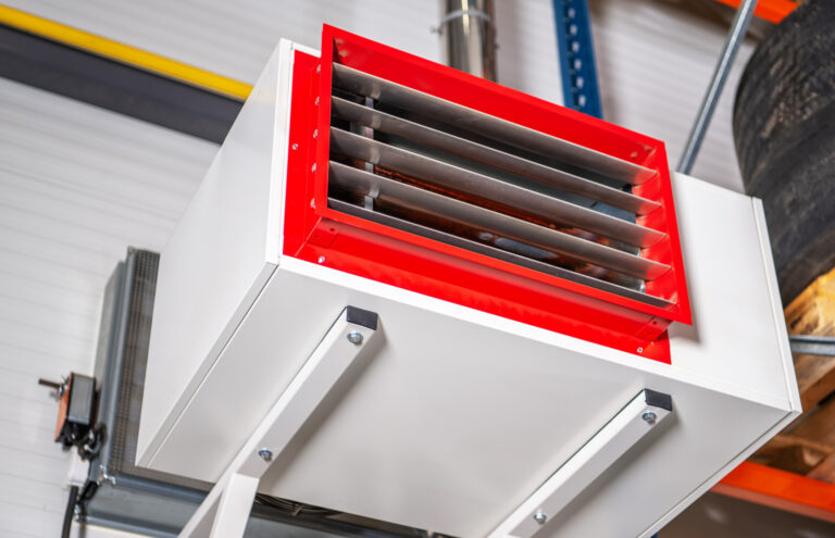 Industrial heater is mounted on a wall in a warehouse setting. The heater has a metal grate and is attached to a metal frame.